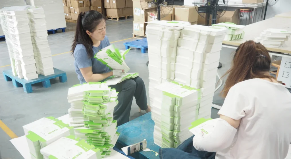 Workers manually assembling and organizing packaging boxes in a factory setting.